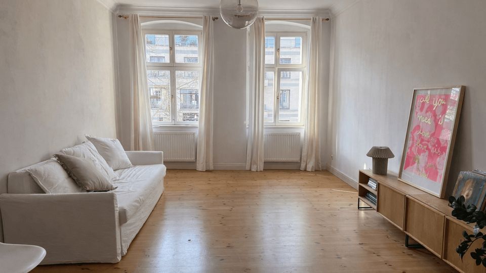 Bright, minimalist living room with light wood floors, a white sofa along one wall, and a low wooden media console on the opposite side. Two tall windows with sheer curtains and radiators beneath them face the street, with a glass globe pendant light hanging from the ceiling.