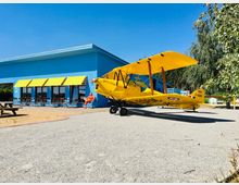 Ein gelbes Doppeldecker-Flugzeug steht auf einem geschotterten Platz vor einem blauen Gebäude mit gelben Markisen. Im Hintergrund sind Sitzplätze vor dem Gebäude und Bäume zu sehen.