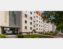 Mehrstöckiges Wohngebäude mit heller Fassade und kleinen Fenstern, umgeben von Rasenflächen, Bäumen und einem gepflasterten Gehweg. Im Vordergrund stehen Fahrräder, und ein Durchgang führt zu einem Innenhof.