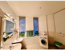 Badezimmer mit zwei Waschbecken, großem Spiegel, Fenster mit grauen Rollläden, Waschmaschine und wandhängender Toilette. Dekorative Elemente wie Pflanzen und Handtücher auf der Fensterbank.