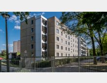 Mehrstöckiges Wohngebäude im Plattenbaustil mit einer Fassade aus strukturierten Betonplatten, flankiert von Bäumen und Begrünung. Im Vordergrund steht ein Metallzaun, dahinter eine kleine Straße und ein Gehweg.