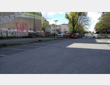 Ein leerer Parkplatz mit gepflastertem Boden, umgeben von Gebäuden, teils mit Graffiti bedeckt, und hohen Bäumen im Hintergrund. Im hinteren Bereich stehen ein weißer und ein roter Transporter sowie Werbetafeln an einem Bauzaun.