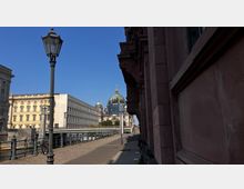 Fußweg entlang eines Flusses mit Geländer und Straßenlaternen, im Hintergrund der Berliner Dom mit seiner grünen Kuppel. Links sind ein schmuckvolles gelbes Gebäude und ein moderner, hellgrauer Bau zu sehen, rechts eine dunkelrote Fassade.