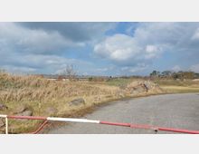 Weite Landschaft mit trockenen Gräsern, verstreuten Felsen und einer teilweise sichtbaren, flachen Industriebebauung im Hintergrund. Im Vordergrund eine Asphaltstraße mit einer rot-weißen Schranke, darüber ein bewölkter Himmel.
