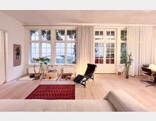 Wohnraum mit großen Fenstern und Glastüren, die auf einen begrünten Außenbereich blicken, ausgestattet mit hellem Holzboden, weißen Vorhängen, einem roten Teppich, einem schwarzen Liegestuhl und minimalistischen Dekorationen wie Pflanzen und Sitzkissen.