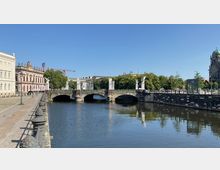 Brücke mit mehreren Bögen und Statuen darauf über einen Fluss, umgeben von historischen Gebäuden und Bäumen; rechts ist ein U-Bahn-Eingang und dahinter eine große Kuppelkirche sichtbar. Der Himmel ist klar und blau.