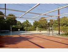 Ein eingezäunter Sport- und Spielplatz mit einem kleinen Fußballtor auf rotem Bodenbelag, umgeben von Gitterstreben und Bäumen im Hintergrund. Rechts im Bild befindet sich ein Spielbereich mit Klettergerüst und Rutsche auf hellem Sandboden.