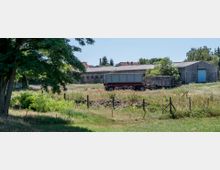 Ein ländlicher Hof mit grauen, flachen Gebäuden und Wellblechdächern, im Vordergrund ein geparkter Anhänger und eine umzäunte, teils bewachsene Wiese. Links ist ein großer Baum zu sehen, der Schatten auf die Wiese wirft.