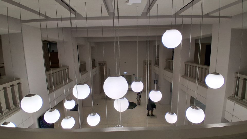 Blick in ein zweigeschossiges, helles Atrium mit umlaufenden Galerien und balustradenartigen Brüstungen an den Seiten. Von der hohen Decke hängen zahlreiche runde Kugelleuchten in unterschiedlichen Größen an langen Kabeln in den offenen Raum.