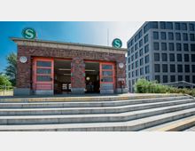 Backsteingebäude mit der Aufschrift "Nordbahnhof" und zwei geöffneten Eingängen, darüber das grüne "S"-Logo für S-Bahn. Im Vordergrund führen Stufen zum Eingang, rechts befinden sich moderne Bürogebäude und grüne Pflanzen.