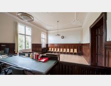 Ein historischer Raum mit Holzvertäfelung an den Wänden, großen Fenstern mit Blick auf Bäume und einer geschmückten Decke. Im Vordergrund steht ein Schreibtisch mit Schreibutensilien, im hinteren Bereich befinden sich Holzstühle in einer Reihe und eine Wanduhr.