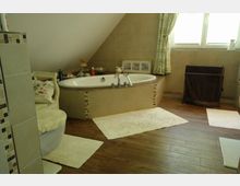 Badezimmer unter einer Dachschräge mit einer eingelassenen Badewanne in Natursteinoptik, hellem Fliesenbelag an den Wänden und dunklem Holzfußboden. Vor der Badewanne liegen helle Teppiche, ein Fenster mit Vorhängen erhellt den Raum.