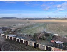 Weitläufige Landschaft mit Feldern und Wiesen, im Vordergrund eine Reihe alter Garagen mit verwitterten Toren. Im Hintergrund sind Industrieanlagen mit rauchenden Schornsteinen zu sehen, daneben Stromleitungen und vereinzelt Bäume.