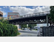 Eine erhöhte Eisenbahnbrücke aus Metall, auf der eine gelbe Bahn fährt, überspannt eine Straße mit parkenden und fahrenden Autos. Im Hintergrund befinden sich ein rotes Backsteingebäude, Bäume und Werbeschilder, umgeben von einem teils bewachsenen und urbanen Umfeld.