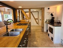 Moderne Küche in einem Fachwerkhaus mit Holzbalken und Fliesenboden. Eine lange Arbeitsplatte aus Holz mit eingebauter Spüle und Gaskochfeld ist zentral, daneben befinden sich ein Holzofen und eine Kaffeemaschine. Im Hintergrund sind ein heller Wohnbereich und große Fenster zu erkennen.