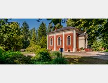 Rotes Backsteingebäude mit weißen Säulenelementen und Rundbogenfenstern, umgeben von einer Gartenlandschaft mit Bäumen, Sträuchern und Blumen. Ein schmaler Weg führt am Gebäude vorbei, das in einer grünen Parklandschaft liegt.