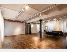 Ein geräumiger, industriell gestalteter Innenraum mit hellen Vorhängen, freiliegenden Stahlträgern und einer abgenutzten Betonbodenfläche. Im Hintergrund sind ein schwarzes Ledersofa, ein Tisch sowie eine weiße Wand mit Fotografiehintergründen erkennbar.