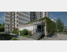 Mehrstöckiges Wohngebäude aus Beton mit horizontal gestreifter Fassade und roten Fensterakzenten. Im Vordergrund ein kleiner Eingangsbereich mit gläsernem Vorbau, einer Treppe und Begrünung, umgeben von Gehwegen und Bäumen.