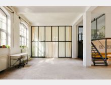 Heller, loftartiger Innenraum mit hohen Decken, großen Sprossenfenstern und einer schwarzen Glas-Trennwand mit Vorhängen. Rechts führt eine Metalltreppe zu einer Galerie mit Glasgeländer; links steht ein schlichter Arbeitstisch an der Fensterfront.