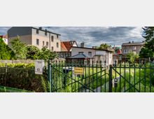 Eine Wohnsiedlung mit einem grünen Metallzaun im Vordergrund, auf dem ein Schild mit der Aufschrift „Einfahrt freihalten!“ hängt. Dahinter befinden sich ein Garten mit hohem Gras, ein kleines Gebäude mit Vordach und mehrstöckige Wohnhäuser im Hintergrund.