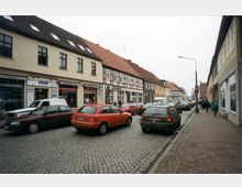 Städtische Straßenszene mit Kopfsteinpflaster, flankiert von zweistöckigen Gebäuden, darunter Fachwerkhäuser und moderne Geschäfte; mehrere parkende und fahrende Autos sowie Fußgänger auf dem Bürgersteig.