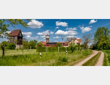 Ländliche Szenerie mit einem unbefestigten Weg, der an einer Wiese und einem bewachsenen Zaun entlangführt. Im Hintergrund befinden sich eine alte Holzscheune, ein Wasserturm und mehrere Gebäude mit roten Ziegeldächern, umgeben von Bäumen und einer weitläufigen Grünfläche unter einem blauen Himmel mit weißen Wolken.