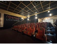 Kinosaal mit leicht ansteigenden Sitzreihen aus roten Polstersitzen, einem Schriftzug „Venus Lichtspiele“ an der dunklen Wand und einer sichtbaren Theke im Hintergrund. Die Decke hat ein gitterartiges Holzdesign, und der Raum ist warm beleuchtet.