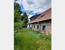 Ein Ziegelhaus mit rotem Satteldach und blauen Fenstern, umgeben von einer verwilderten Wiese und Bäumen. Im Hintergrund sind ein Nadelbaum und teils bewölkter Himmel zu sehen.