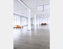 Großer, heller Raum mit glatten grauen Böden, weißen Wänden und mehreren weißen Säulen, die die Fläche gliedern. An den Seiten befinden sich vereinzelte Sitzmöbel wie ein gelbes Sofa und ein brauner Sessel.