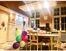 Heller Raum mit hohen Decken, großen Sprossenfenstern und warmen Wandfarben. In der Mitte steht ein Esstisch mit Stühlen, dekoriert mit Ballons und Geschenken, während im Hintergrund eine antike Kommode und weiße Türen zu sehen sind.