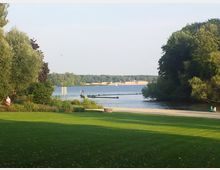 Ein See mit angrenzendem Park, umgeben von Bäumen und Wiesenflächen. Im Vordergrund ist eine gepflegte Rasenfläche, während am Ufer Spaziergänger und kleinere Bootsstege zu sehen sind.