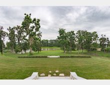 Blick von einer Steintreppe auf eine weitläufige Parklandschaft mit gepflegtem Rasen, hohen Bäumen und einem kleinen Teich im Hintergrund. Der Himmel ist bedeckt, und die Szenerie wirkt ruhig und offen.