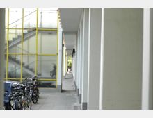 Überdachter Durchgang mit einer Reihe von Pfeilern rechts und einer wandhohen Fensterstruktur mit gelben Rahmen links; mehrere Fahrräder sind entlang der Fenster abgestellt. Im Hintergrund ist eine Treppe hinter den Fenstern sichtbar.