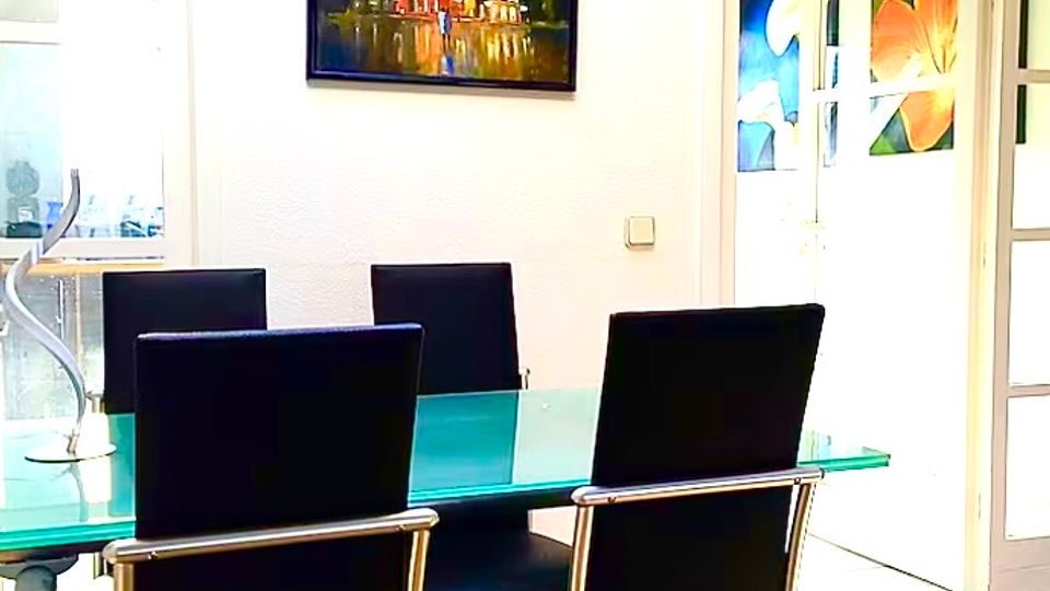 Heller Essbereich mit rechteckigem Glastisch und mehreren schwarzen Hochlehner-Stühlen auf Holzboden. An der weißen Wand hängt ein gerahmtes Bild, daneben führen Glastüren in angrenzende Räume mit farbigen Wandbildern.