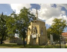 Historische Backsteinkirche mit Spitzbogenfenstern und einem schlanken Kirchturm, umgeben von Bäumen und einzelnen Gebäuden im Hintergrund; eine Wiese und ein Bauzaun im Vordergrund.