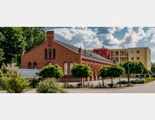 Backsteingebäude mit gotischen Fenstern und Satteldach, daneben moderne gelbe und rote Wohngebäude im Hintergrund. Im Vordergrund befinden sich ein Parkplatz mit vereinzelten Bäumen und niedriger Vegetation.