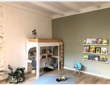 Kinderzimmer mit Hochbett aus Holz, integriertem Regal für Bücher und Spielzeug sowie einer kleinen Holzleiter. An der olivgrünen Wand hängen weiße Wandregale mit Kinderbüchern, daneben steht ein blauer Kinderstuhl. Der Fußboden ist aus hellem Holz, und eine Zimmerpflanze sowie Spielsachen dekorieren den Raum.