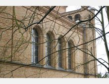 Backsteinfassade einer Kirche mit hohen Rundbogenfenstern und verzierten Glasarbeiten, teilweise von kahlen Ästen verdeckt. Im Hintergrund ist ein kirchlicher Turm mit Arkaden zu erkennen.