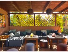 Überdachte Terrasse mit Holzdecke, gemütlicher Sitzecke mit grauen Polstern und bunten Kissen, mehreren Holztischen und Hockern. Im Hintergrund ein Gitter mit rankenden Pflanzen, das von sonnendurchflutetem Laub umrahmt ist.