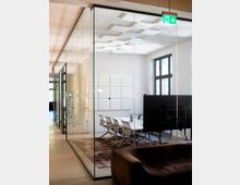 Moderner Konferenzraum in einem Büro, durch eine raumhohe Glaswand vom Flur getrennt. Im Raum stehen ein langer Tisch mit mehreren Stühlen, große Fenster, eine Decke mit quadratischen Akustikpaneelen sowie ein grünes Notausgangsschild über der Tür.