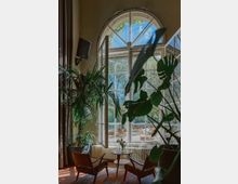 Ein gemütlicher Innenraum mit zwei Sesseln und einem kleinen runden Tisch, umgeben von großen Zimmerpflanzen. Im Hintergrund ein großes, halbkreisförmiges Fenster mit Blick auf eine sonnige Terrasse mit Sitzmöbeln und einen Garten.