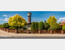 Ein gepflegter Garten mit Zierbäumen und Sträuchern, eingefasst von einem Zaun aus Holz und Backsteinpfosten, im Hintergrund ein runder Wasserturm aus Backstein. Die Szene liegt an einer Straße unter blauem Himmel mit wenigen Wolken.