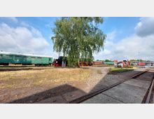 Historische Eisenbahnszene mit einer grünen Waggonreihe und einer schwarzen Dampflokomotive im Hintergrund, die teilweise von einem großen Baum verdeckt wird. Im Vordergrund befinden sich Kopfsteinpflasterwege und Schienen, rechts ein ausgestelltes Paar rot-weißer Eisenbahnräder.