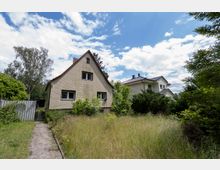Blick auf ein freistehendes Einfamilienhaus mit steilem Satteldach und hell verklinkerter Fassade in einer grünen Wohngegend. Davor liegt ein verwilderter Garten mit hohem Gras, Büschen und einem schmalen Weg; im Hintergrund steht ein weiteres Haus unter blauem Himmel mit Wolken.