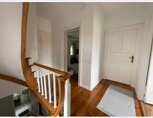 Heller Hausflur mit weiß gestrichenen Wänden, einer Holztreppe mit weißen Geländerstäben und Dielenboden. Drei geschlossene weiße Türen, ein kleiner grauer Teppich und ein offener Durchgang führen zu weiteren Räumen.