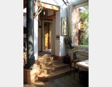 Heller Innenraum mit einem kleinen Holzofen, einer Treppe aus Holz und einem großen Fenster mit Blick auf einen Garten. Der Raum ist teils dekoriert mit Kunstgegenständen, darunter eine Wandskulptur und Vasen, und wird von natürlichem Licht durchflutet.