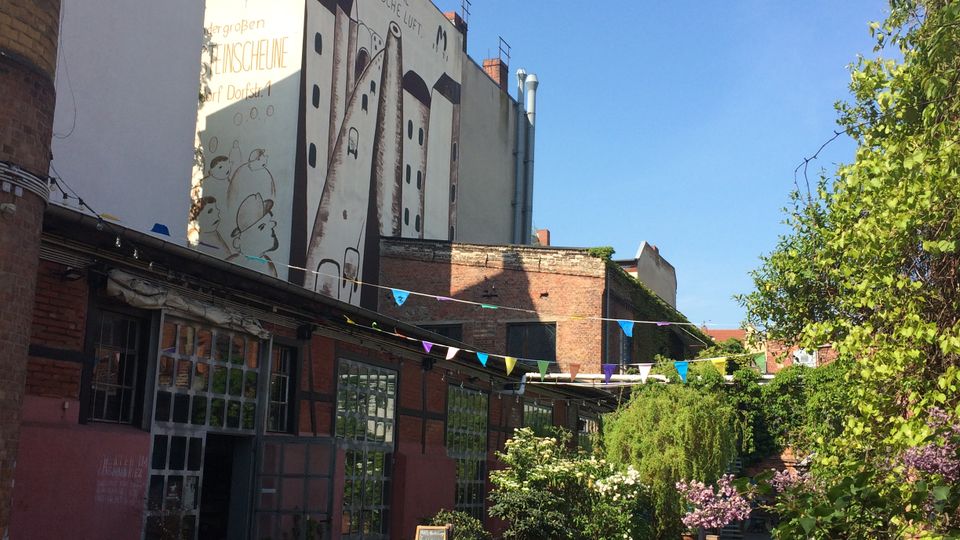Innenhof in einem umgebauten Industrieareal mit Backstein- und Werkstattgebäuden, großen Fensterfronten und einer bemalten Giebelwand im Hintergrund. Über dem gepflasterten Weg hängen bunte Wimpelketten, daneben stehen Tische und Stühle sowie üppige Grünpflanzen und blühende Sträucher.