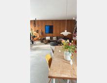 Wohnzimmer mit Holzvertäfelung an der Wand, einer dunklen Couch mit Kissen, weißen und schwarzen Poufs, Zimmerpflanzen und einem abstrakten blauen Bild. Im Vordergrund ein Esstisch mit Holzplatte, gelben und weißen Stühlen sowie einer Vase mit bunten Tulpen.