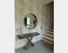 Moderner Flur mit grauer, strukturierter Wand, einem runden Spiegel, einer schmalen Konsole mit dunkler Glasoberfläche und einer goldfarbenen Schlangenfigur. Rechts führen Holzstufen eine Treppe hinauf.