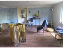 Ein schlicht eingerichteter Raum mit blauen Wänden und Holzboden. Im Vordergrund steht ein Gestell mit bunten Stoffen, daneben sind verschiedene Möbelstücke wie Stühle, ein kleiner Tisch, eine Stehlampe und dekorative Objekte wie ein Sonnenschirm und Vasen sichtbar.